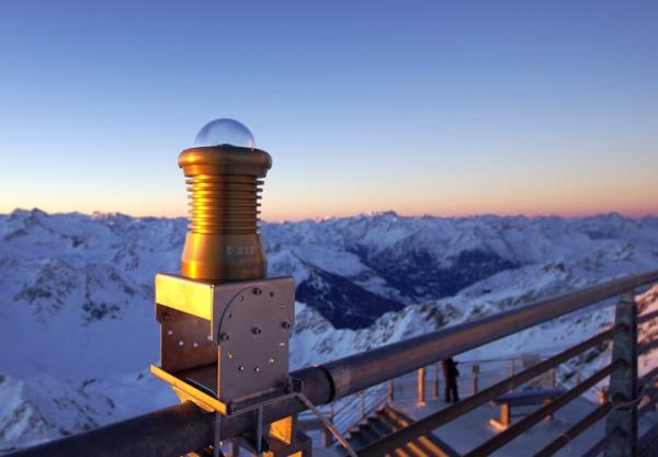 A camera, which looks like a glass sphere, attached to a me<em></em>tal stand on a railing overlooking mountains.